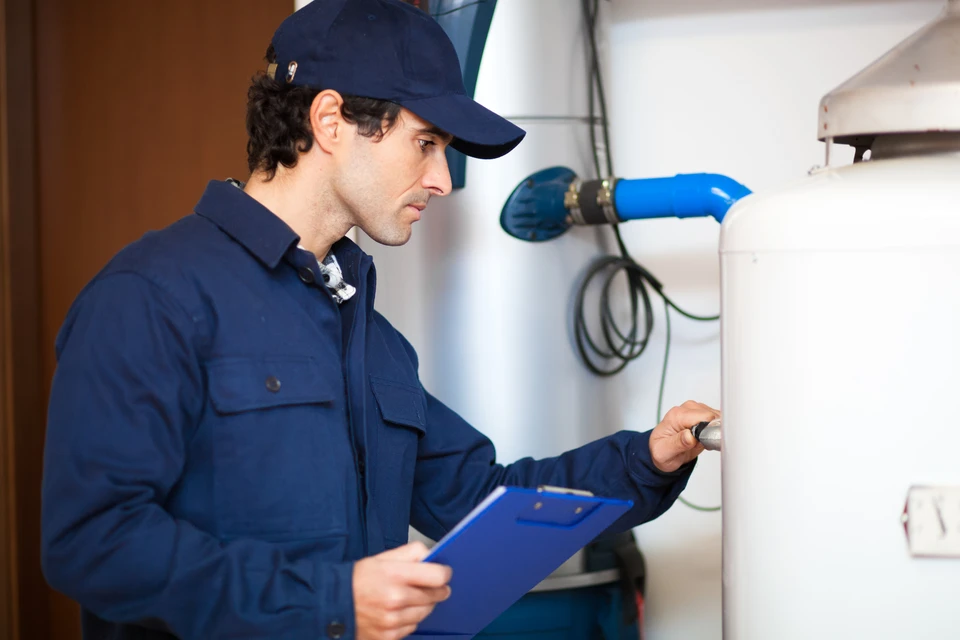 Technicien en uniforme bleu et casquette effectuant la maintenance d'un chauffe-eau électrique dans un local technique résidentiel